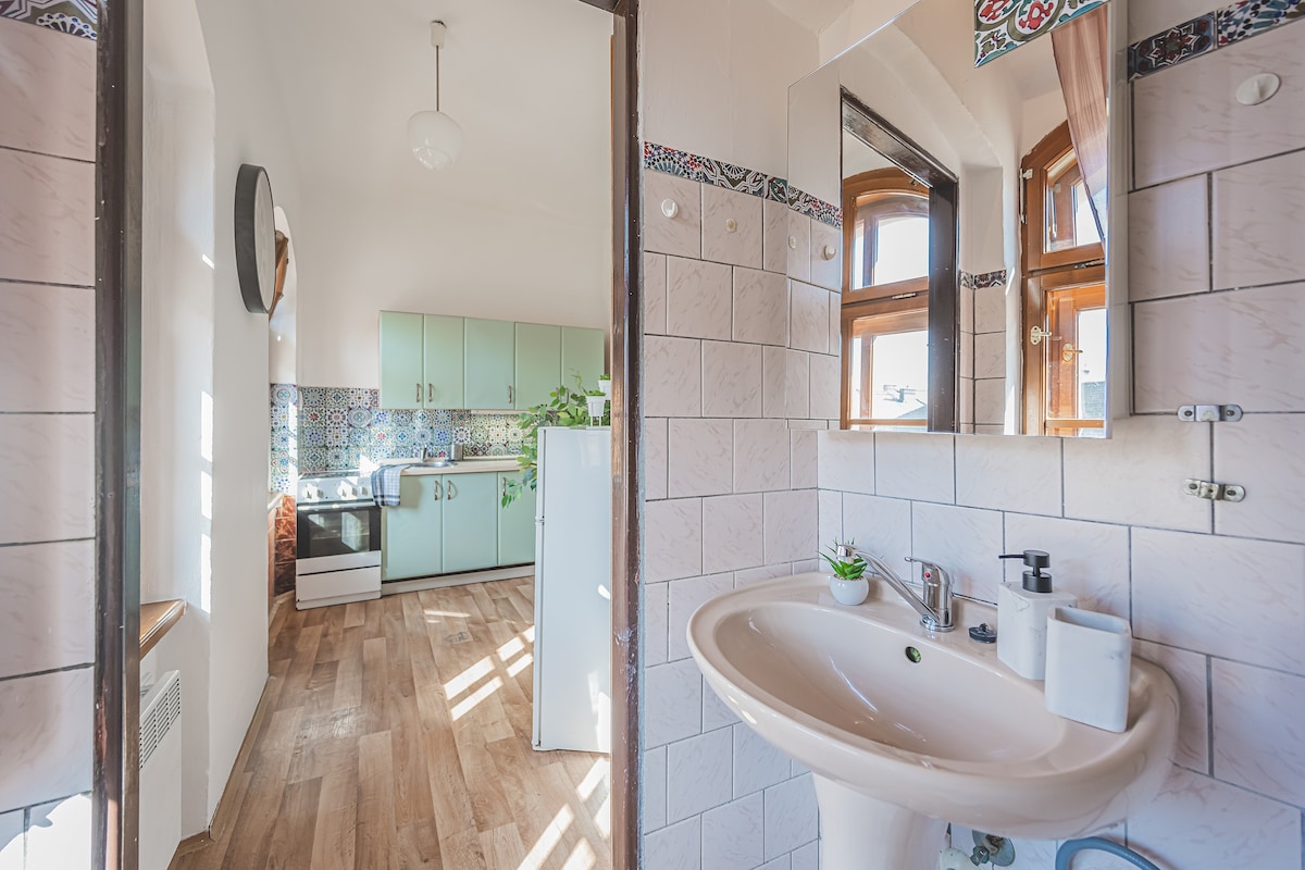 A bright bathroom features a classic sink with a mirror above it. Natural light enters through windows, illuminating the tiled walls. The doorway opens to a modern kitchen area with light blue cabinetry and colorful backsplash tiles, enhancing the airy feel of the space.