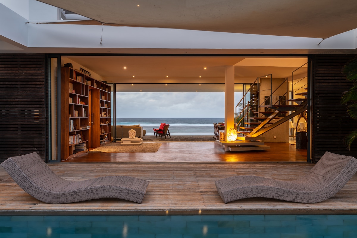 A spacious living area is seen through large sliding glass doors, featuring an open design that connects to a wooden sundeck. Two lounge chairs rest by a tiled pool, while a collection of books fills the nearby shelves. Cozy seating faces the ocean view.