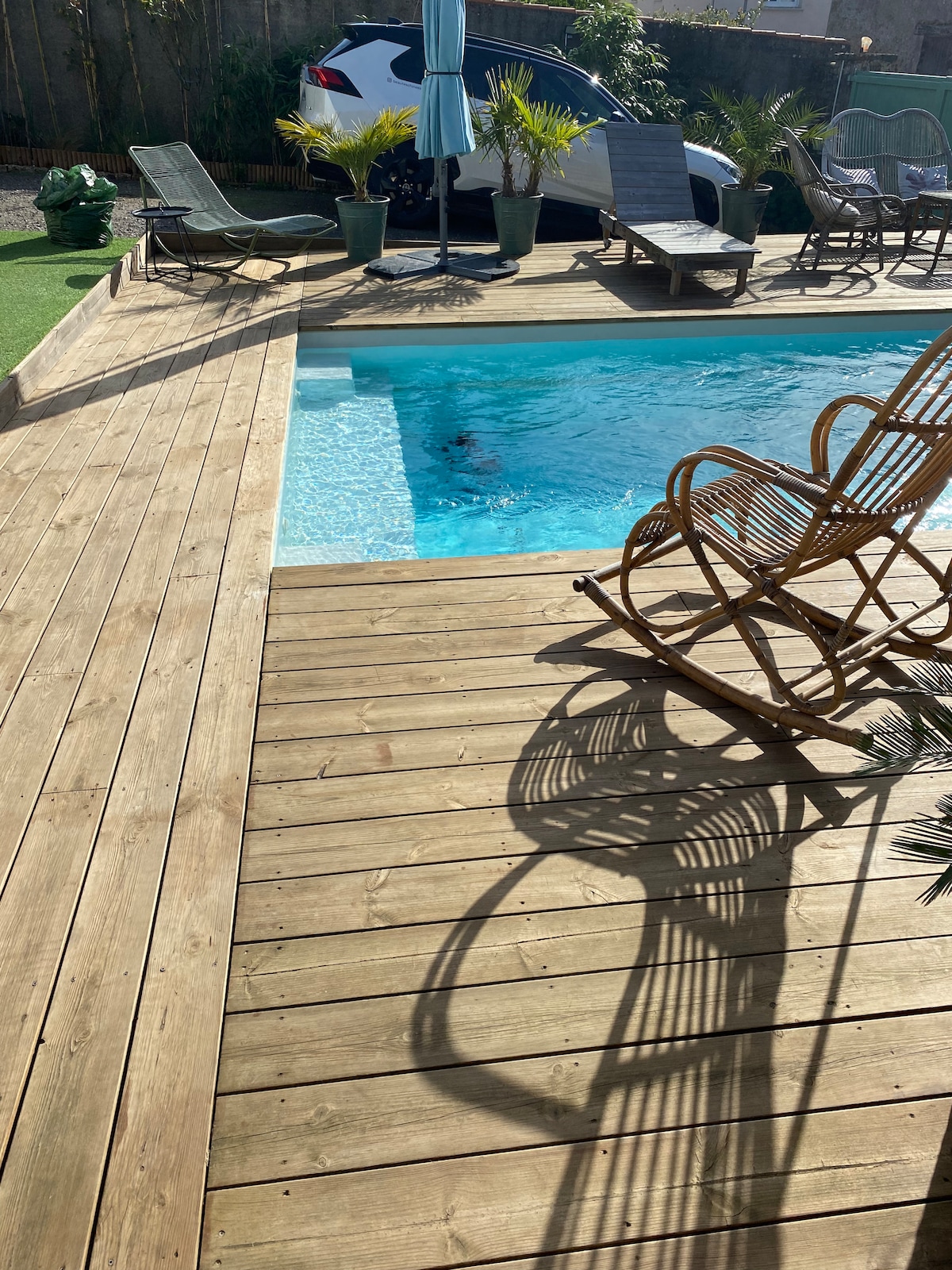A wooden deck surrounds a clear blue swimming pool, with several lounge chairs positioned nearby. A tall umbrella provides shade, and lush plants add a natural touch. The reflection of the chairs creates shadows on the deck, contributing to the tranquil atmosphere.