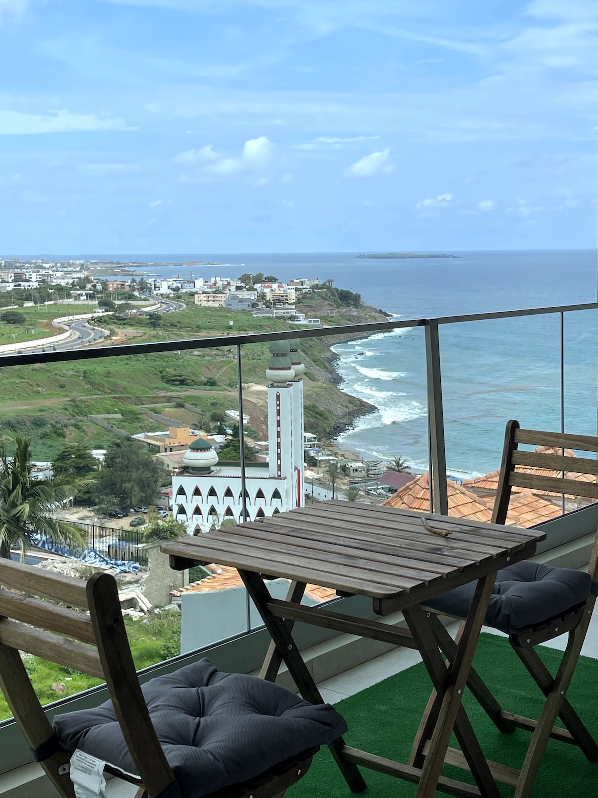 A balcony features a small wooden table and two folding chairs, providing a vantage point of the Atlantic Ocean and coastal landscape. The Mosque of Divinity is visible nearby, surrounded by greenery and residential buildings. The sky is partly cloudy, with coastal cliffs extending in the distance.