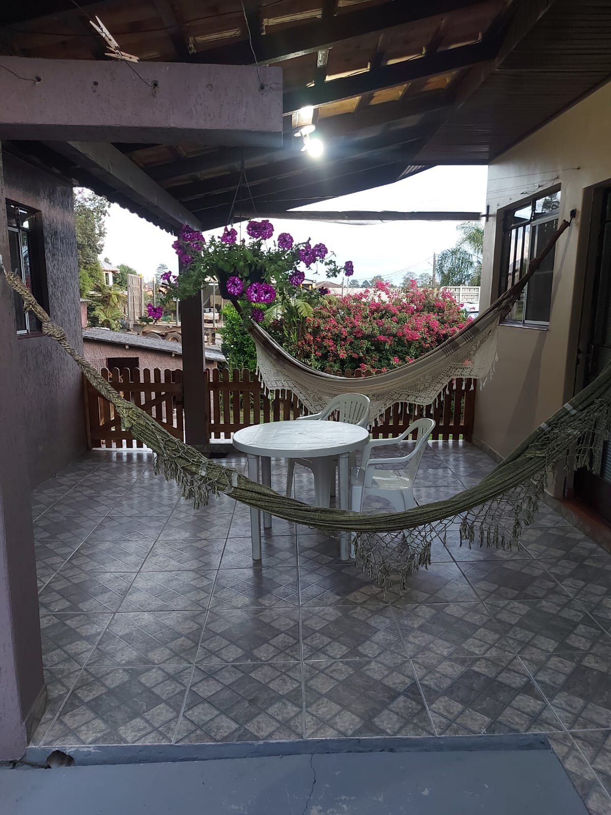 A covered terrace features two hammocks gently swaying between the posts, complemented by a small round table and a white chair. Lush flowers in vibrant hues spill over the balcony railing, enhancing the serene outdoor setting.