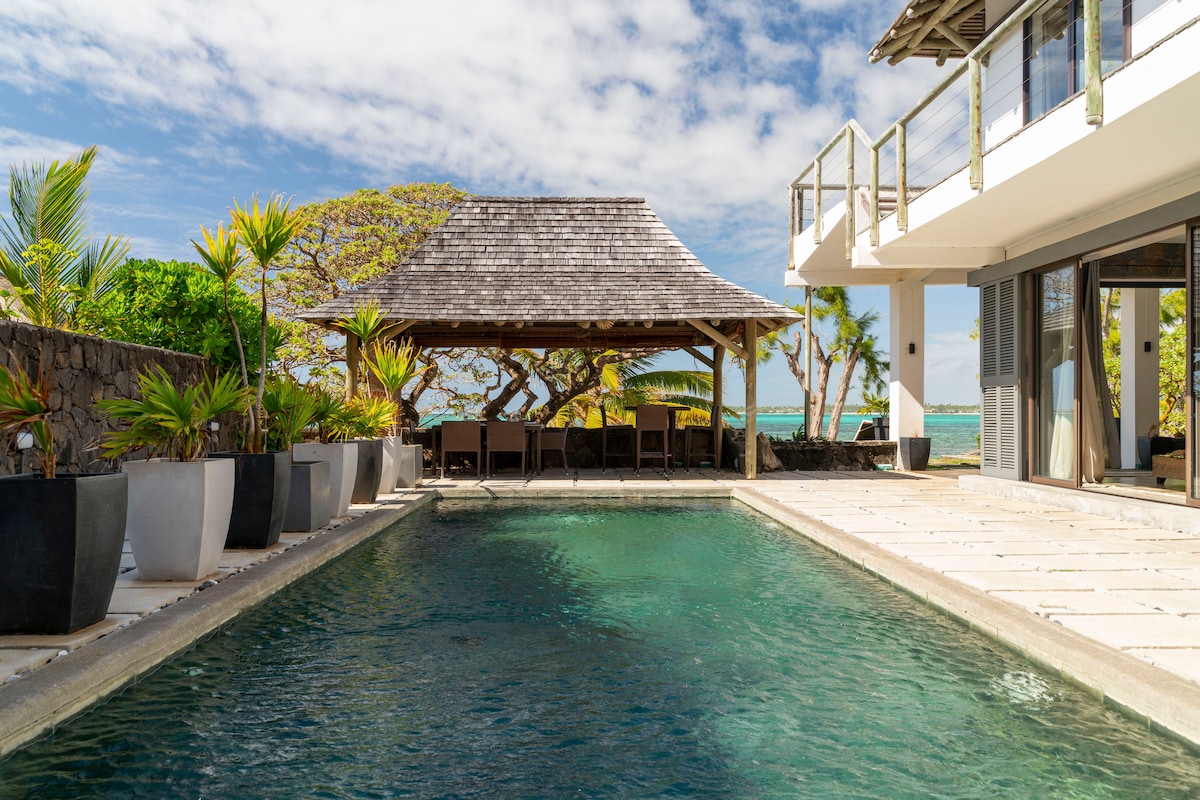 A serene outdoor space features a rectangular swimming pool bordered by stone tiles. A thatched-roof gazebo with dining chairs sits nearby, surrounded by various potted plants. Lush greenery and a glimpse of turquoise water are visible in the background under a partly cloudy sky.