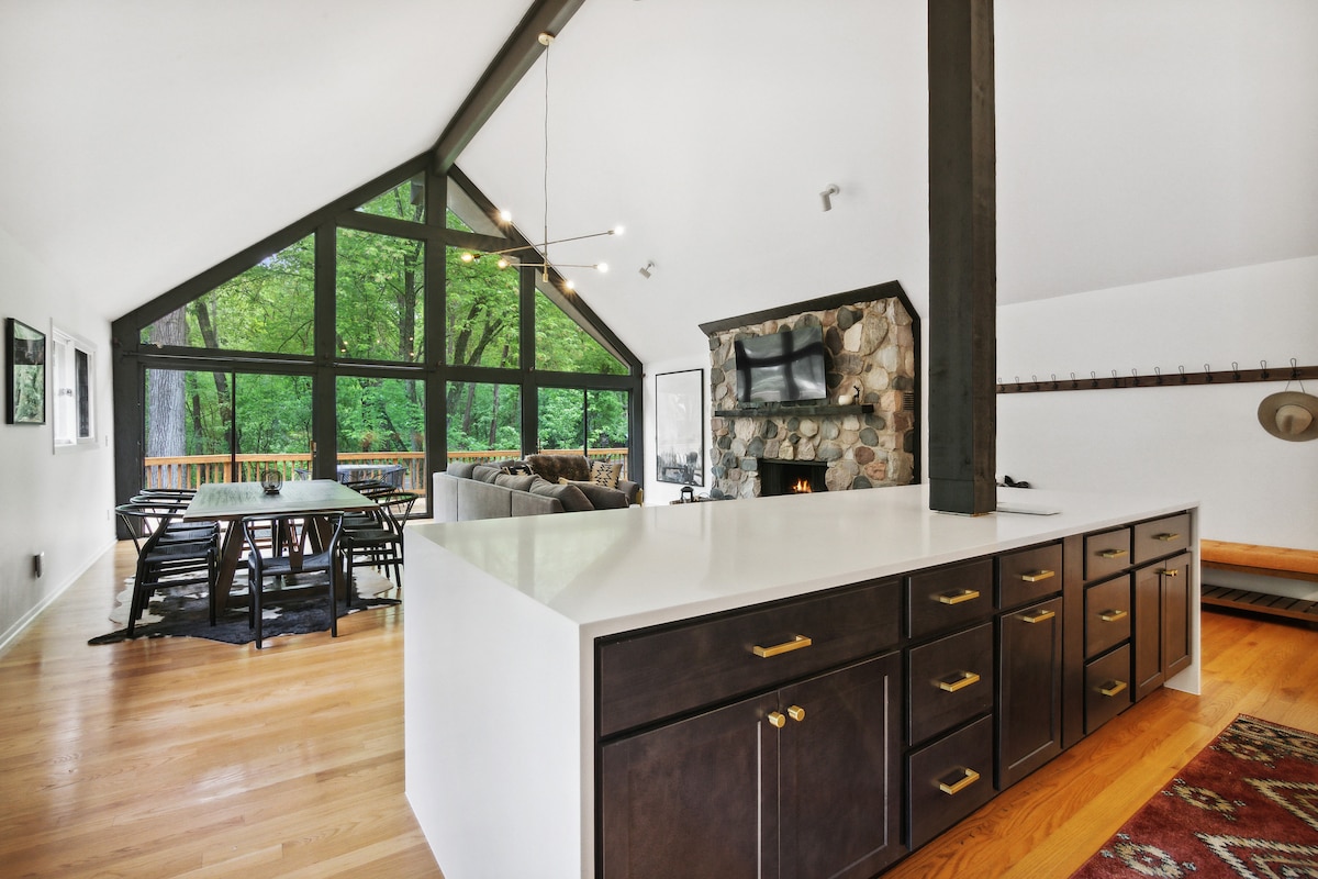 An open living space features a modern kitchen island with dark cabinetry and white countertops. A cozy seating arrangement is visible by a stone fireplace, with large windows offering views of the surrounding trees. Natural light brightens the area, creating an inviting atmosphere.