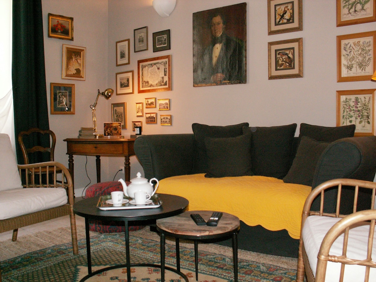 A cozy living area is highlighted by a plush dark sofa adorned with a throw blanket. Two light-colored chairs frame a round coffee table, which holds a teapot and cups. The wall is decorated with a collection of framed art and photographs, adding character to the space.