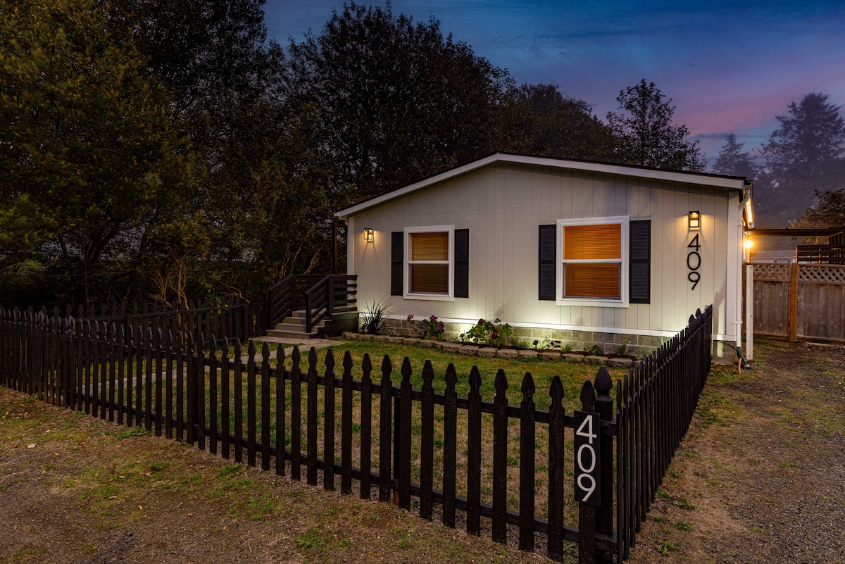 A cozy cottage with a well-maintained yard is surrounded by a decorative black fence. The exterior features light-colored siding and multiple windows, with warm lighting illuminating the entrance. A pathway leads to the front door, inviting guests into a welcoming atmosphere.