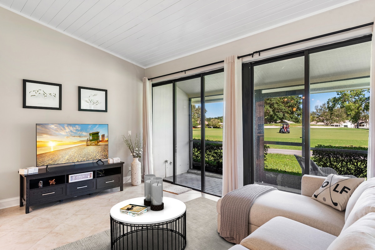A modern living space features a large television atop a sleek black media console. Floor-to-ceiling glass doors open to a patio, showcasing serene golf course views. Natural light fills the room, highlighting a cozy seating area with a light grey throw draped over a white sofa.