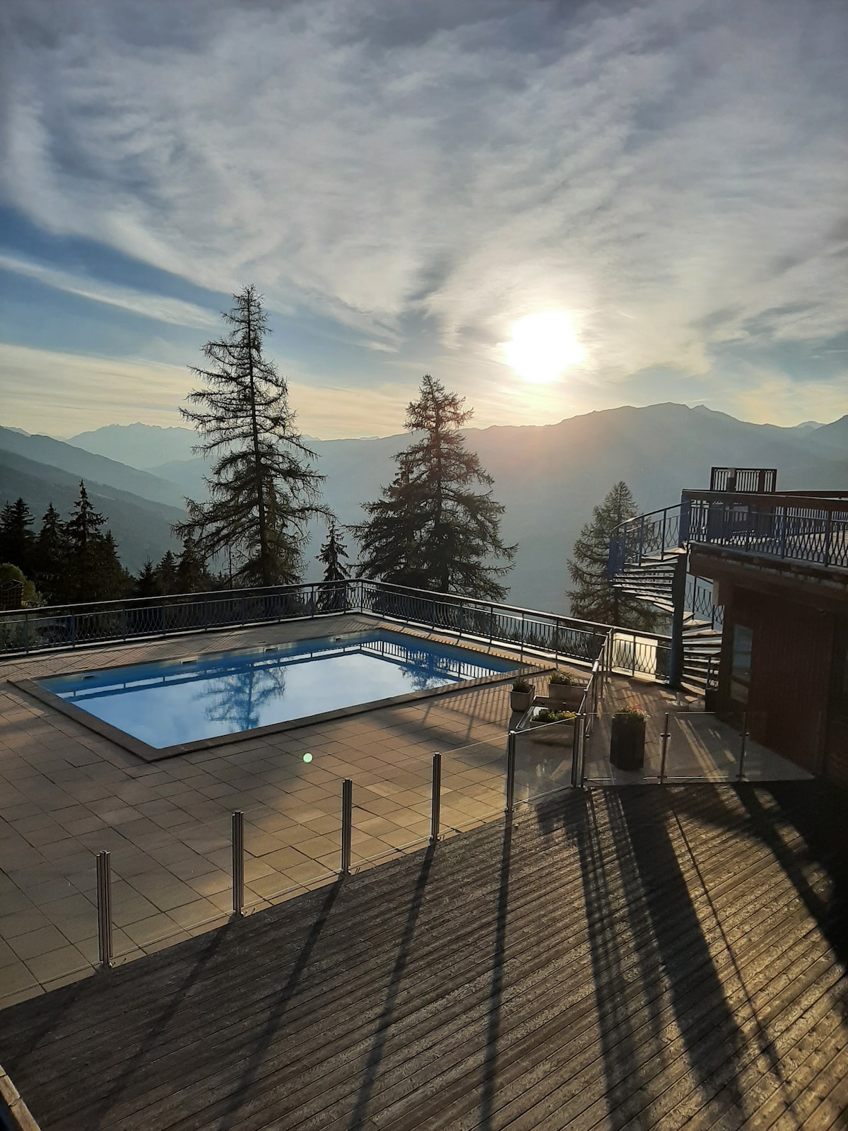 A serene outdoor pool is visible, reflecting the soft light of the setting sun. Surrounding trees create a natural barrier, while the distant mountains provide a picturesque backdrop. A wooden deck extends in the foreground, enhancing the tranquil atmosphere of the space.