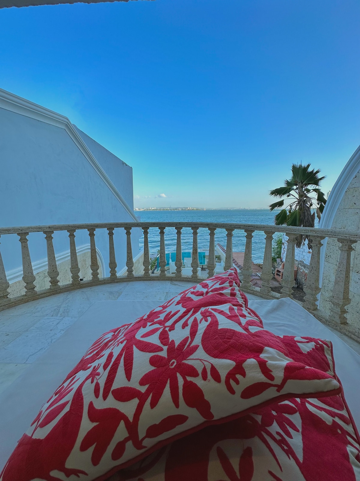 A balcony view is presented, featuring a decorative railing. Bright cushions with floral patterns are arranged on a lounge area, while the serene blue panorama of the bay and palm trees are visible in the background under a clear sky.