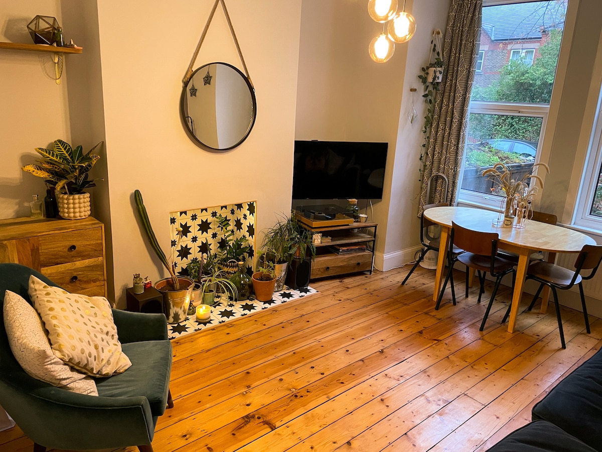 A cozy living space features a green armchair and a wooden console beneath a wall-mounted television. A round dining table with four chairs is positioned nearby. Decorative plants, a mirror, and a patterned rug create an inviting ambiance. Natural light enters through large windows.