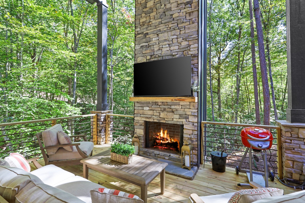 An outdoor patio area showcases a stone fireplace with a fire burning within. Comfortable seating arrangements surround a wooden coffee table, and a modern grill is positioned nearby. Lush greenery is visible in the background, creating a serene and natural setting.