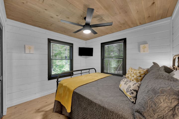 Main floor bedroom with queen bed, barn door, Roku TV, and closet with hangers & luggage rack. There is a ceiling fan and blinds on the two windows. Beautiful wooded views.