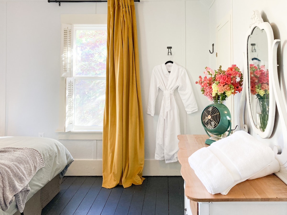 A serene room features a spacious bed dressed in soft linens. A vintage fan sits on a wooden dresser beside neatly folded towels. A golden curtain provides a warm accent, while a bouquet of colorful flowers brightens the space. Sunlight filters in through a nearby window.