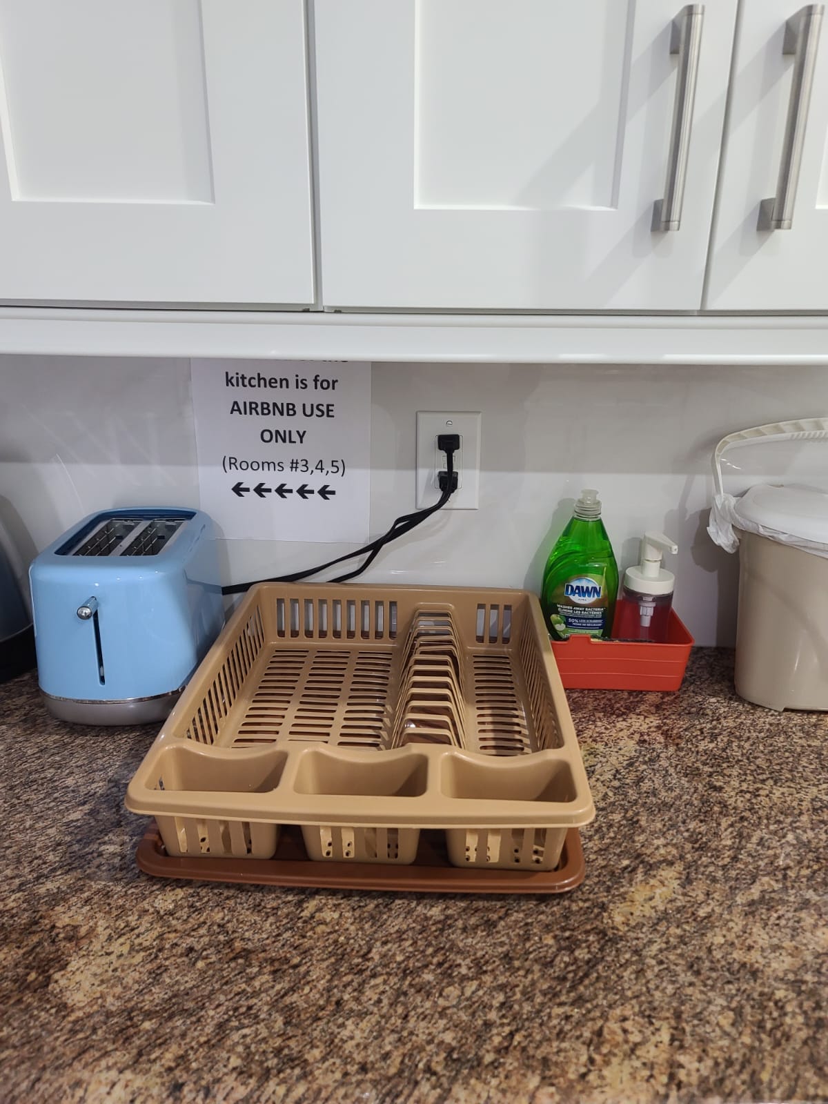 A dedicated drying rack is shown on a granite countertop, accompanied by dish soap and other cleaning supplies. A blue toaster is visible in the background, while a sign indicating kitchen usage for Airbnb guests is mounted on the wall nearby.