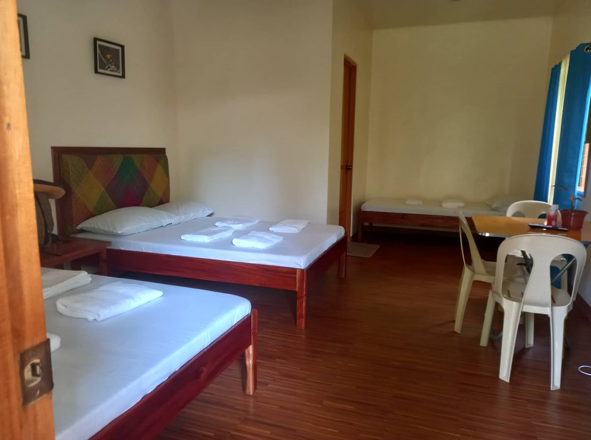 A well-lit room features three beds arranged against the walls, each neatly made with white linens. A small dining area with a wooden table and white chairs is positioned near the window, enhancing the space's functionality.
