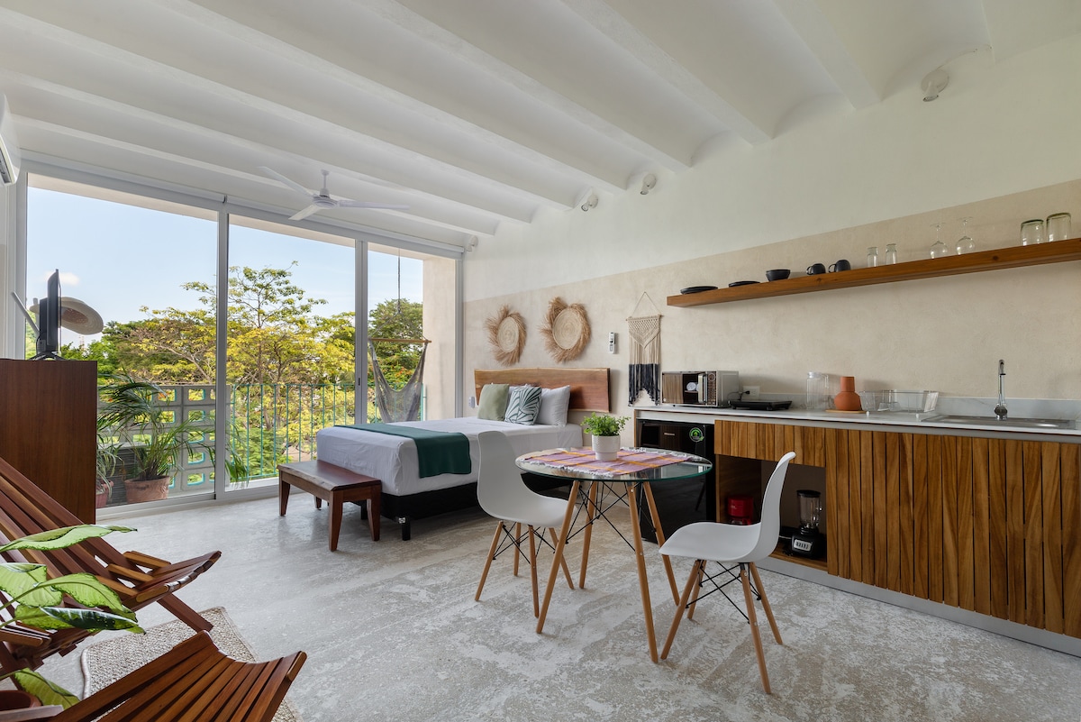A bright and modern studio features a bed positioned against a wall of natural wood grain. A small dining area with white chairs and a round table is situated beside the kitchenette, which includes a sink and microwave. Large glass doors open to a balcony with views of greenery.
