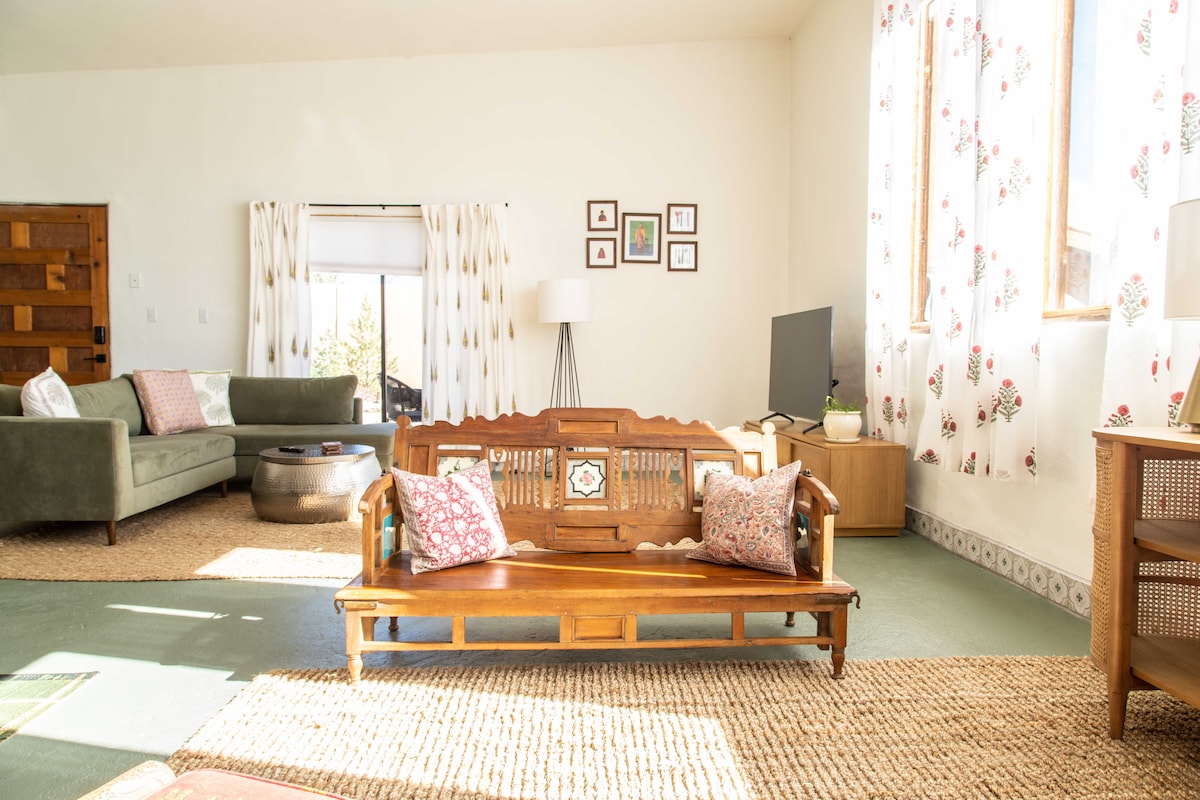 A spacious living area features a traditional wooden bench with decorative pillows, complemented by a green sofa and a media console. Light filters in through large windows adorned with floral curtains, illuminating the textured area rug and enhancing the cozy atmosphere.