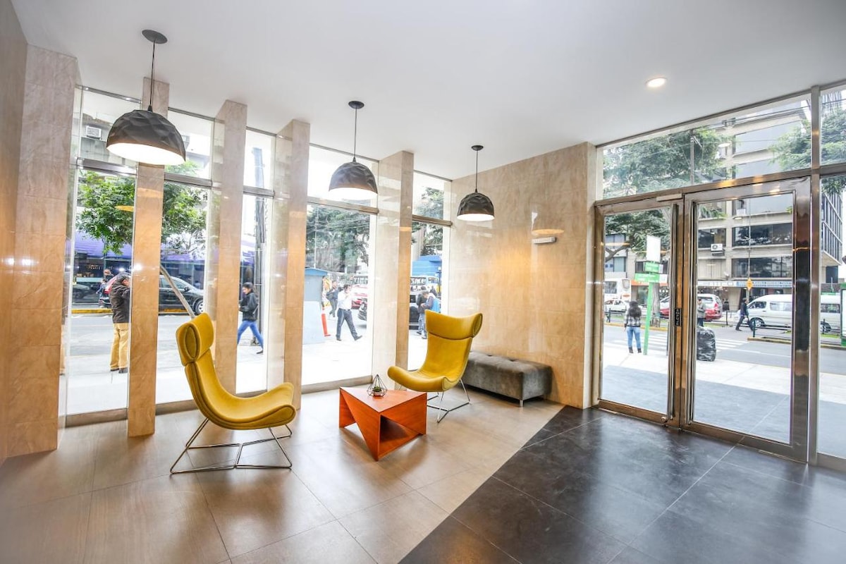 A modern lobby features large floor-to-ceiling windows allowing natural light to fill the space. Two yellow armchairs are positioned around a small orange table. Neutral-colored walls and flooring complement the design. Decorative pendant lights hang from the ceiling, enhancing the inviting atmosphere.