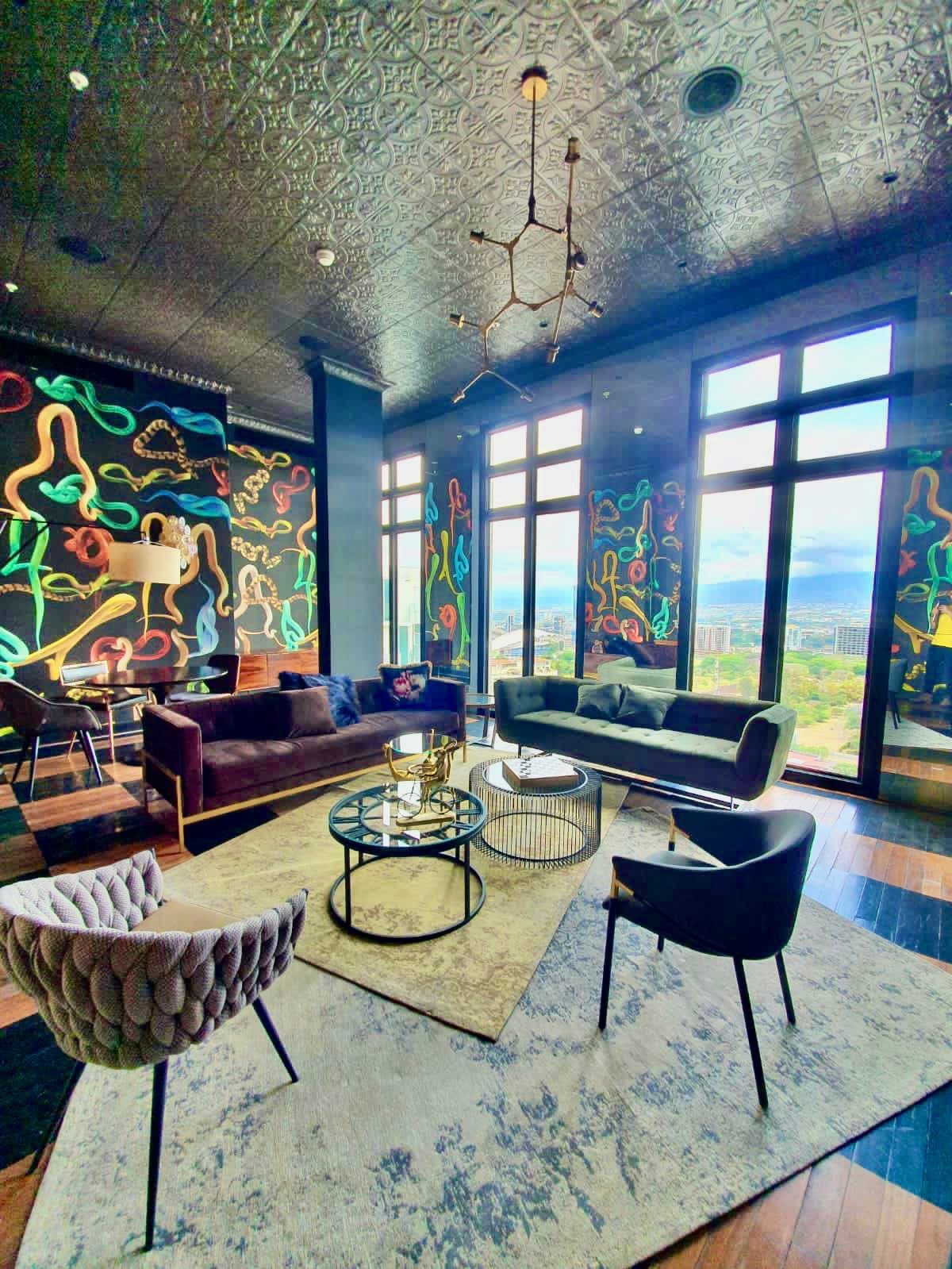 An inviting lounge area features a dark velvet sofa paired with contemporary armchairs. A round coffee table is centered on a large area rug, while large windows frame views of the surrounding landscape. Colorful wall art adds a unique touch to the elegant space.