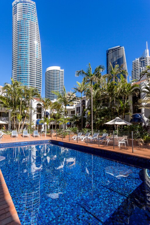 A sparkling blue swimming pool reflects the clear sky, surrounded by lush palm trees and lounge chairs. Towering buildings can be seen in the background, adding a modern touch to the serene outdoor setting.
