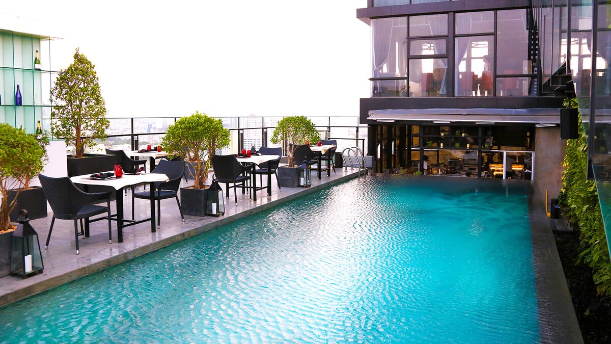 An elevated pool area features a serene turquoise water surface, flanked by dining tables with black chairs. Lush greenery enhances the space, while the modern structure offers an open view of the surroundings, providing a tranquil setting for relaxation.