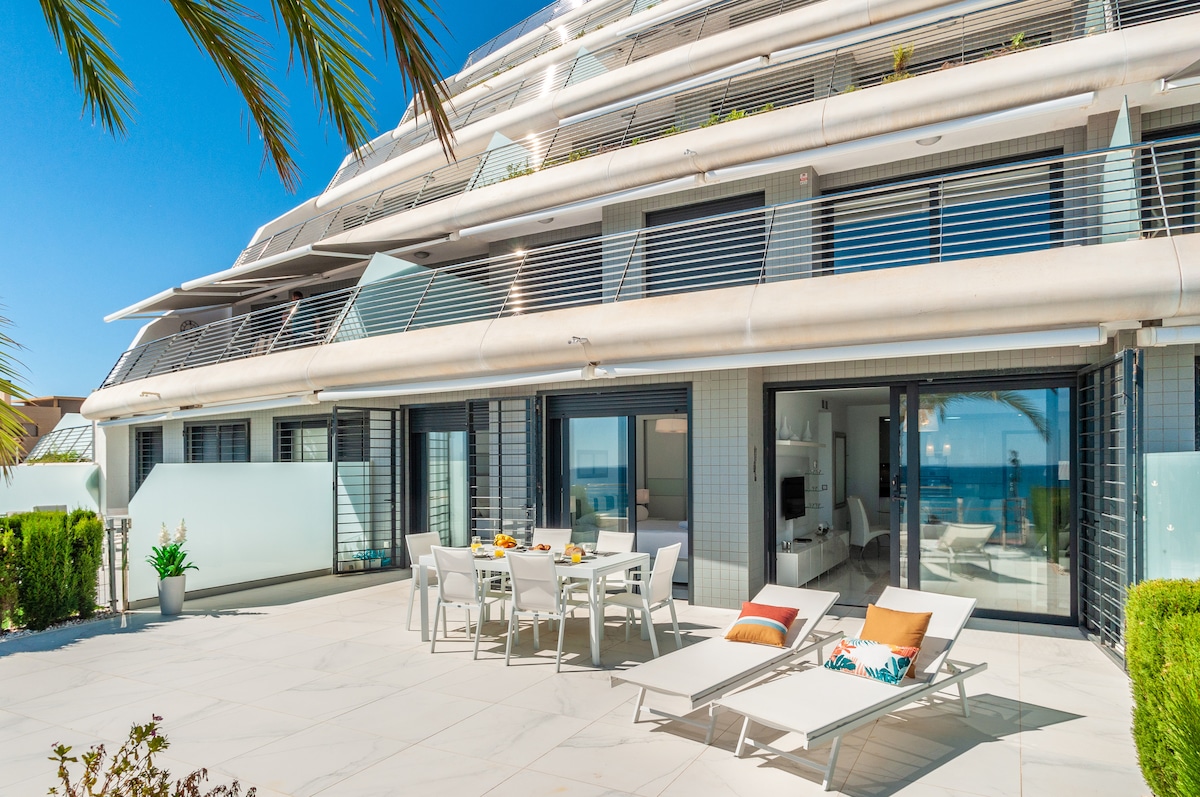 A spacious terrace is showcased, featuring modern outdoor furniture, including a large dining table adorned with tableware. Sun loungers with decorative cushions are positioned for relaxation, while lush greenery and a clear view of the sea enhance the tranquil setting.