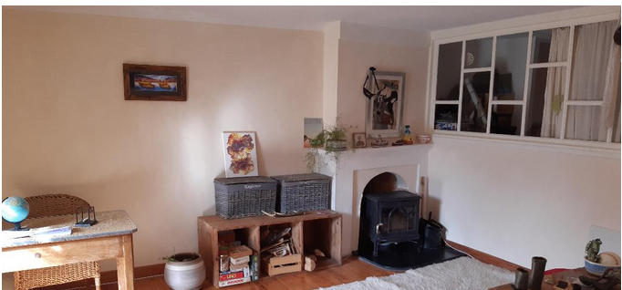 A cozy living area is showcased, featuring a traditional wood-burning stove set against a wall. Natural light filters through a large window, complemented by decorative elements including artwork and a plant. The space includes a woven basket and a rustic wooden table.