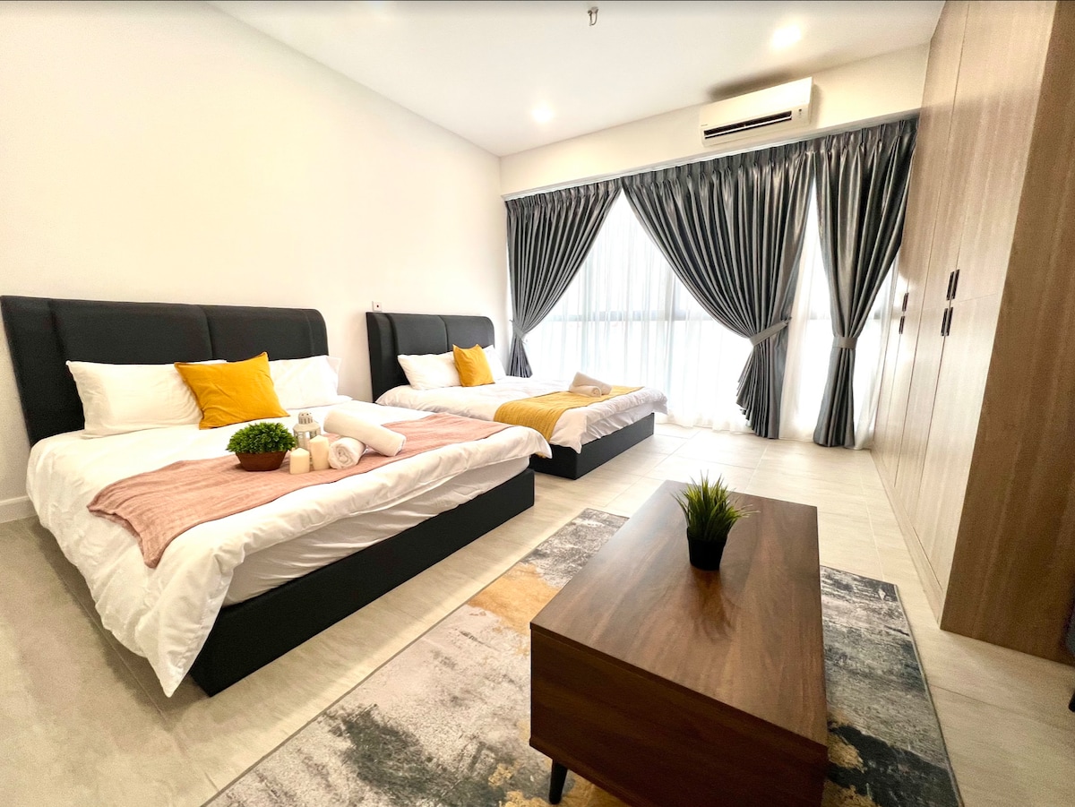 The spacious bedroom features two queen beds, each adorned with soft linens and decorative pillows. A wooden coffee table sits in the center, surrounded by a vibrant area rug. Large windows draped with elegant curtains allow natural light to fill the room.