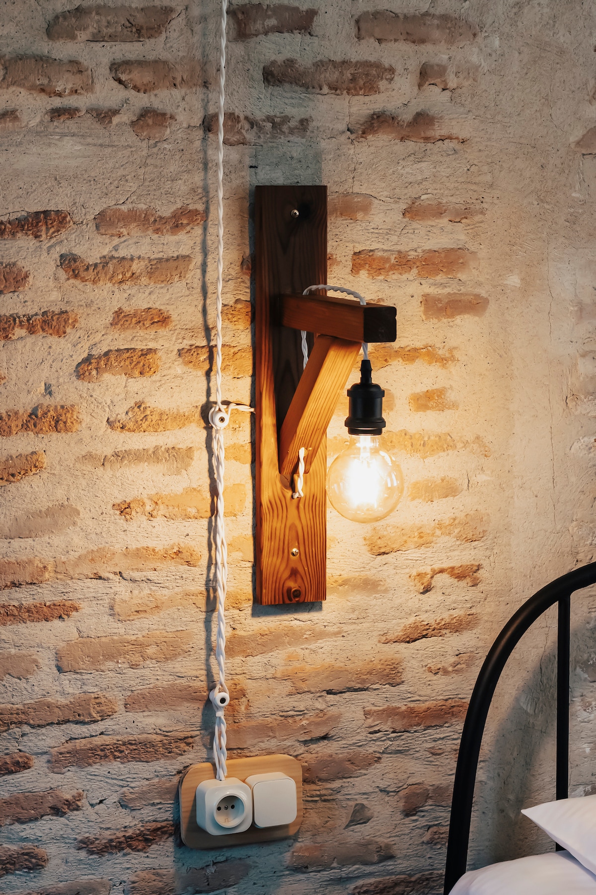 A wall-mounted light fixture is positioned next to the bed, featuring a vintage-style bulb that casts a warm glow. The fixture is attached to a wooden panel against a textured brick wall, enhancing the room's character.