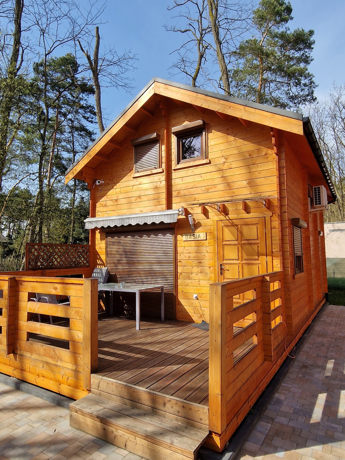 A wooden cabin is shown with a welcoming front deck. The exterior features large windows and a door, surrounded by trees. Outdoor furniture is positioned on the deck, providing a comfortable space to relax. Sunlight streams onto the cabin, enhancing its natural wood finish.