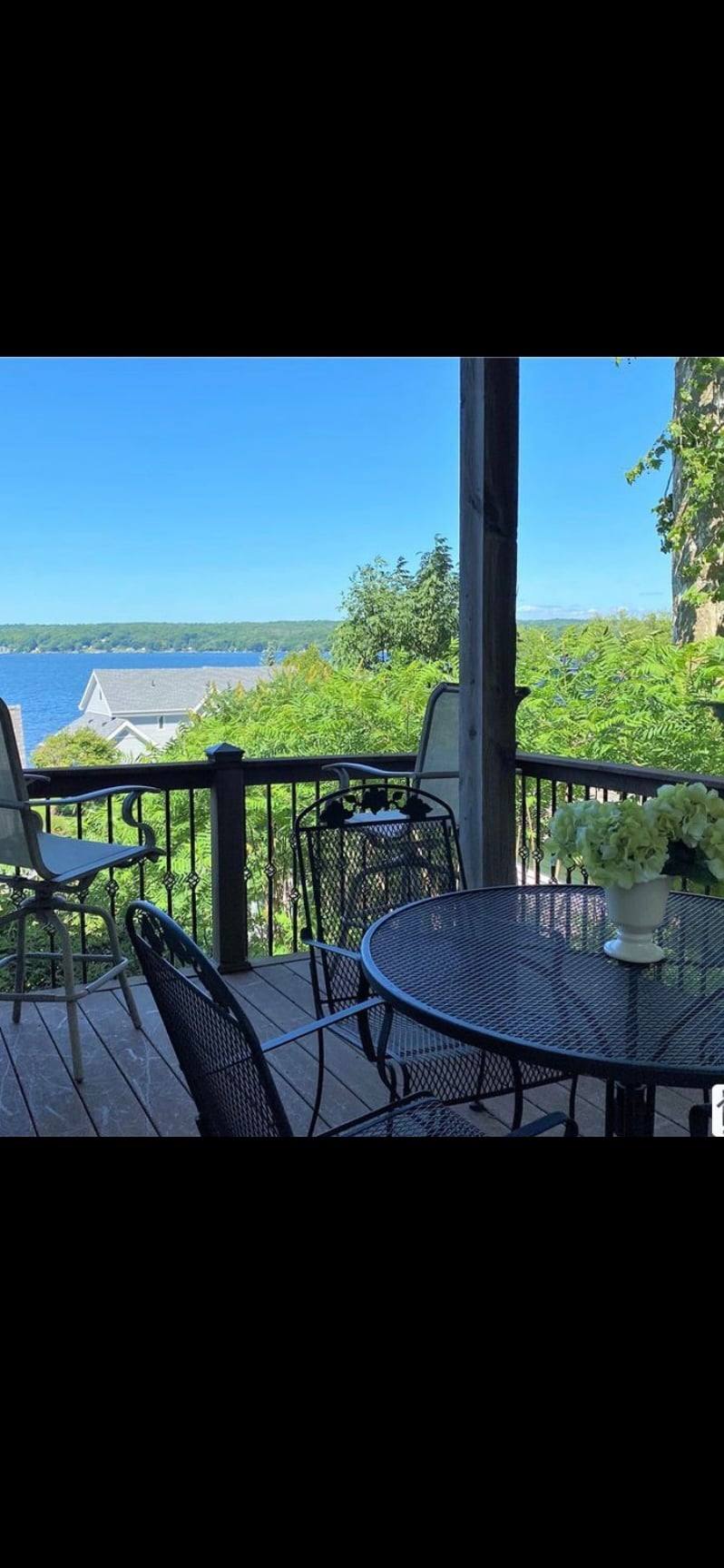 A covered outdoor patio features a black metal table with matching chairs, surrounded by lush greenery. The tranquil view of Chautauqua Lake is visible in the background, under a clear blue sky. A vase with flowers sits on the table.