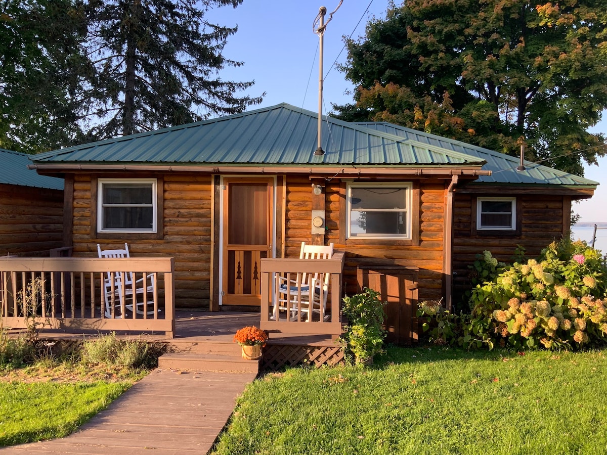 A rustic cabin with a green metal roof features wooden log exterior and a welcoming front porch. White rocking chairs are positioned on the porch, complemented by flower pots. Lush greenery surrounds the cabin, offering a tranquil setting near Oneida Lake.