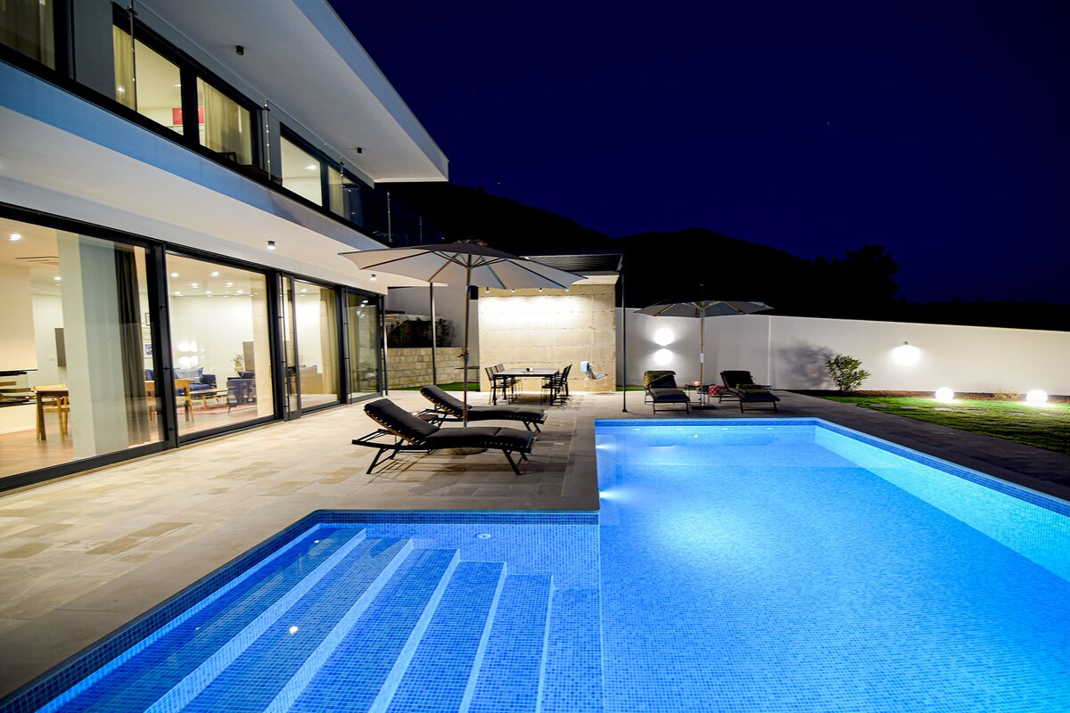 The outdoor area features a sleek private pool illuminated in blue, accompanied by several sun loungers and large umbrellas for shade. Modern glass doors provide a view of the interior, while a dining table is set outside for alfresco meals near the pool.