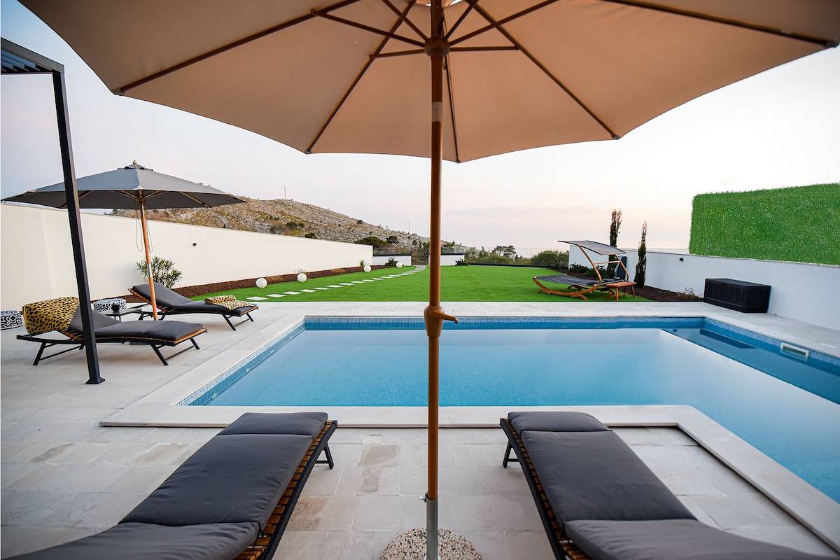 A serene pool area features a clear water pool surrounded by sun loungers and large umbrellas for shade. The green lawn stretches beyond the pool, while a stylish lounge chair and a hammock enhance the outdoor relaxation options.