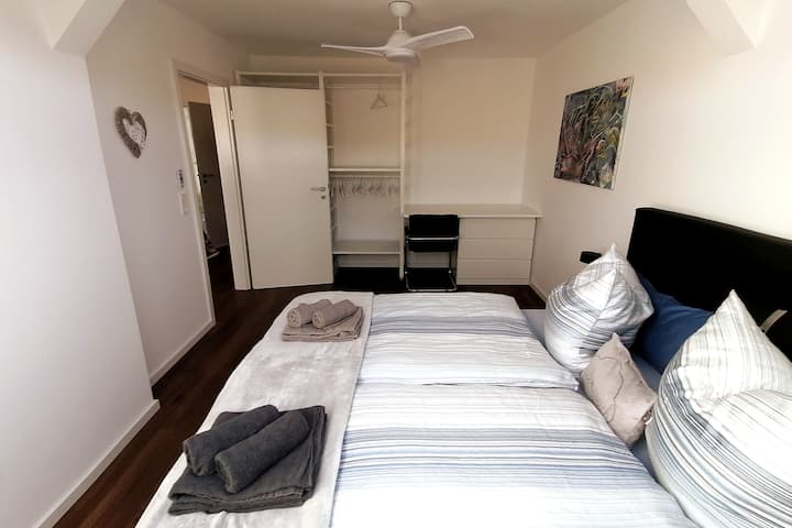 Dormitorio espacioso con cama de muelles de 180 cm x 200 cm: lámparas de postre, enchufes USB, ventilador de techo, cómoda, sistema de almacenamiento para ropa, escritorio pequeño y TV.