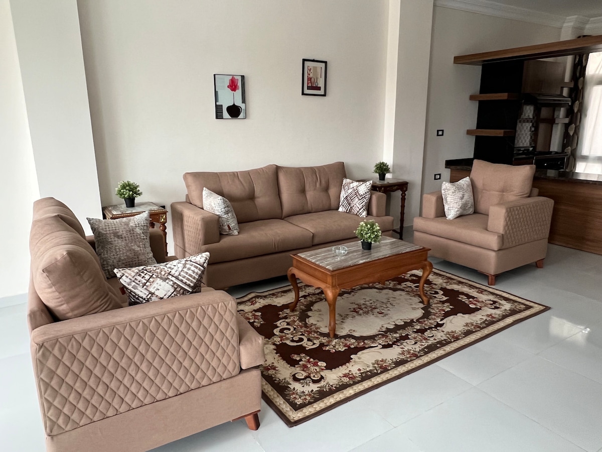 A spacious living area is furnished with a comfortable sofa set and two armchairs, complemented by decorative cushions. A wooden coffee table is placed on an ornate rug, with potted plants adding a touch of greenery. Framed artwork hangs on the wall, enhancing the overall decor.