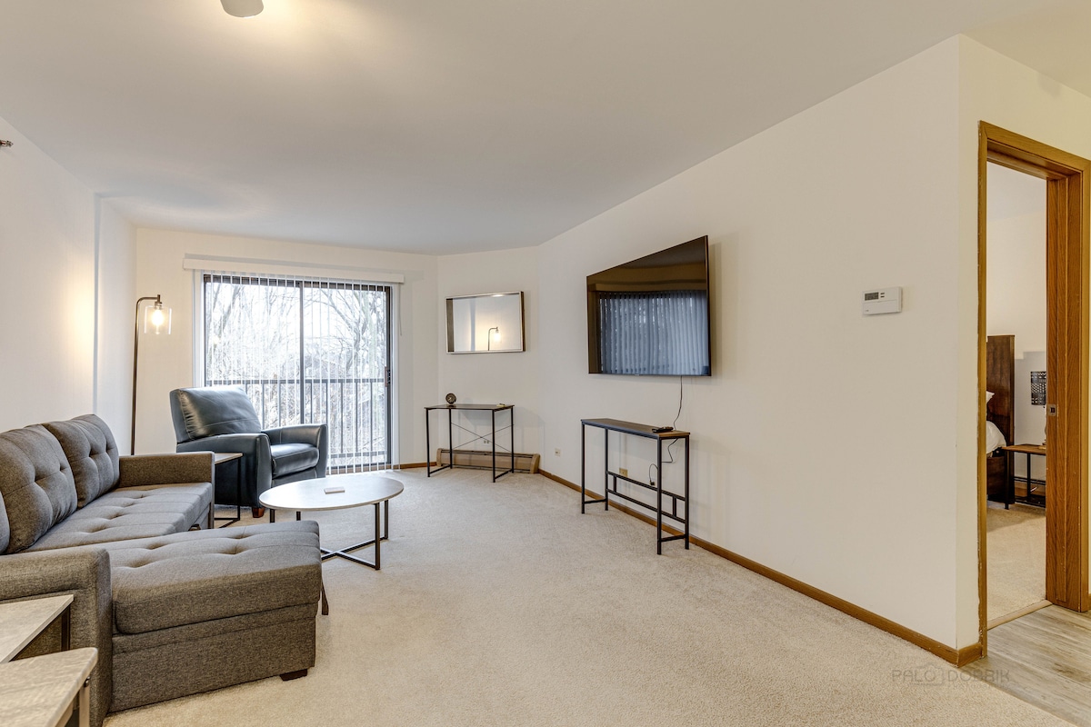A spacious living area showcases a comfortable gray sofa and armchair, paired with a round coffee table. A flat-screen TV is mounted on the wall opposite a large window, allowing natural light to fill the room. A mirror and décor piece enhance the space.