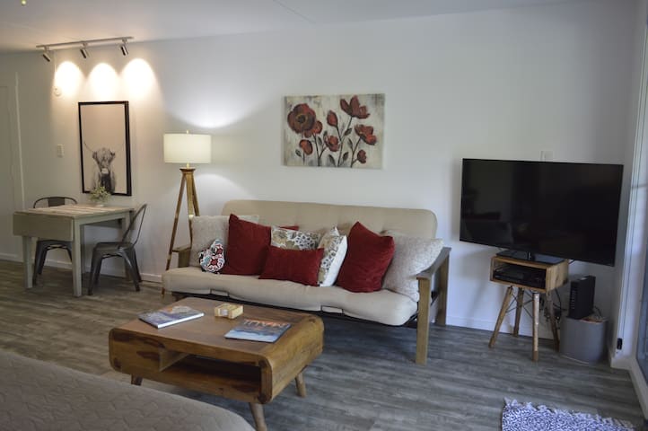 A comfortable seating area features a futon adorned with decorative pillows, paired with a wooden coffee table. A small dining table is positioned nearby, illuminated by a softly glowing lamp. A flat-screen television is mounted on the wall, enhancing the cozy ambiance.