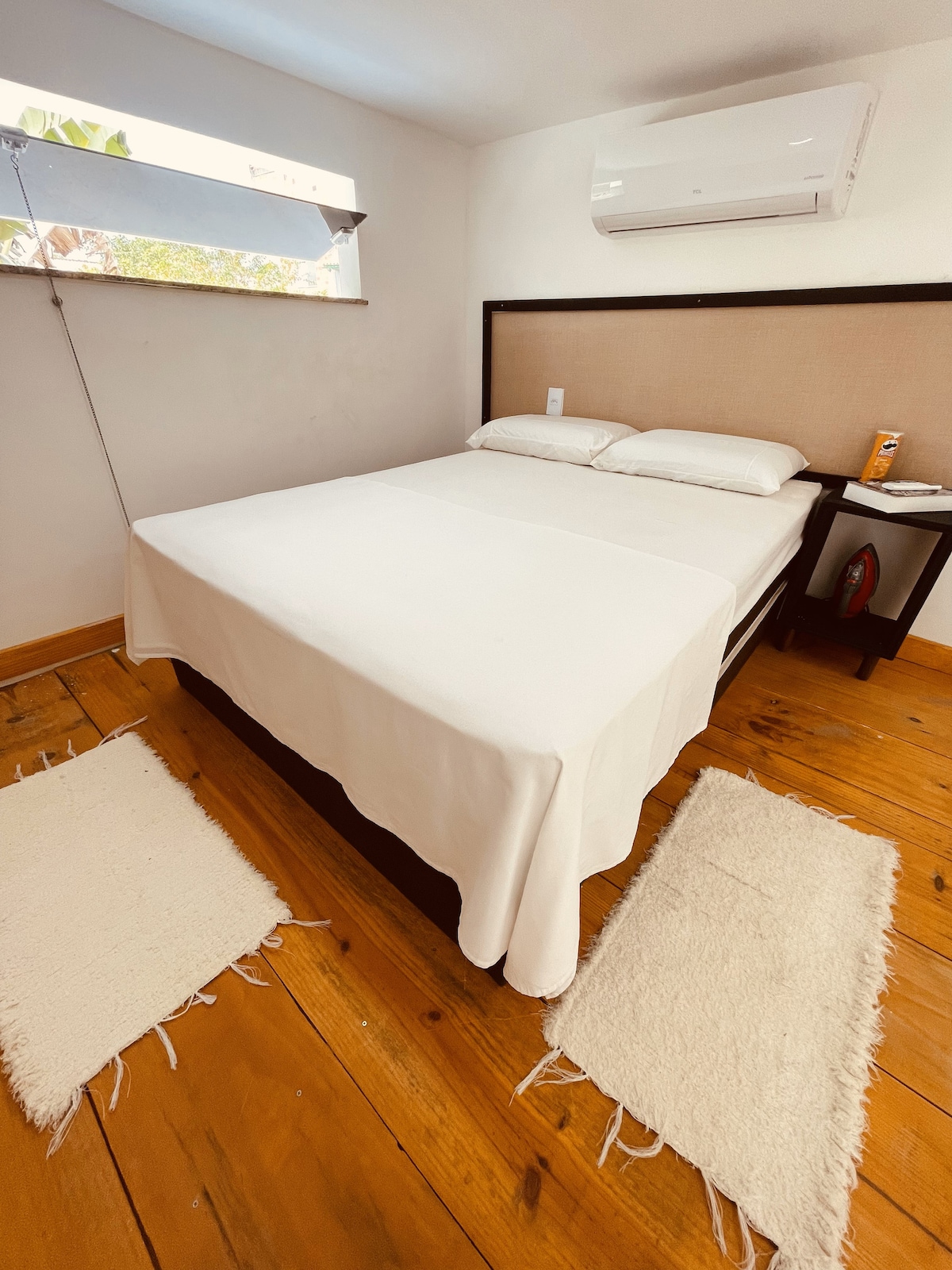 A simple sleeping area is featured with a spacious bed dressed in white linens. Two soft area rugs are placed on the polished wooden floor beside the bed. A window allows natural light to enter, and an air conditioning unit is mounted on the wall.