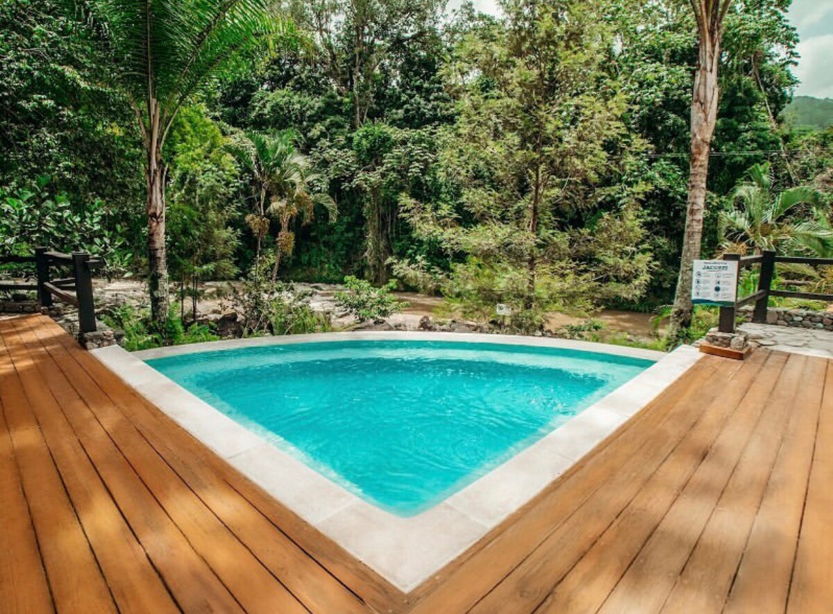 A triangular swimming pool with clear blue water is surrounded by a wooden deck. Lush greenery and trees are visible in the background, creating a natural setting that enhances the tranquil atmosphere.