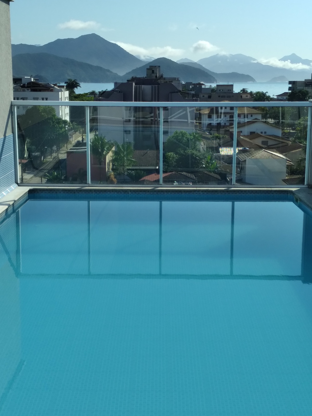 A rooftop pool is showcased, featuring clear, tranquil water surrounded by a glass railing. In the background, mountains rise under a clear sky with gentle clouds, offering a serene view over the surrounding neighborhood.