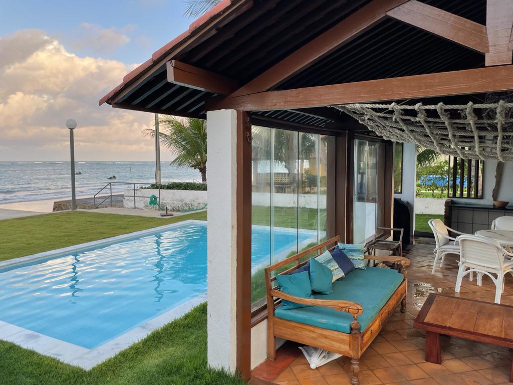 A spacious outdoor area is shown, featuring a private pool with clear water reflecting the sky. Comfortable seating is available under a covered porch, with a direct view of the ocean. Lush grass surrounds the pool, enhancing the beachfront setting.