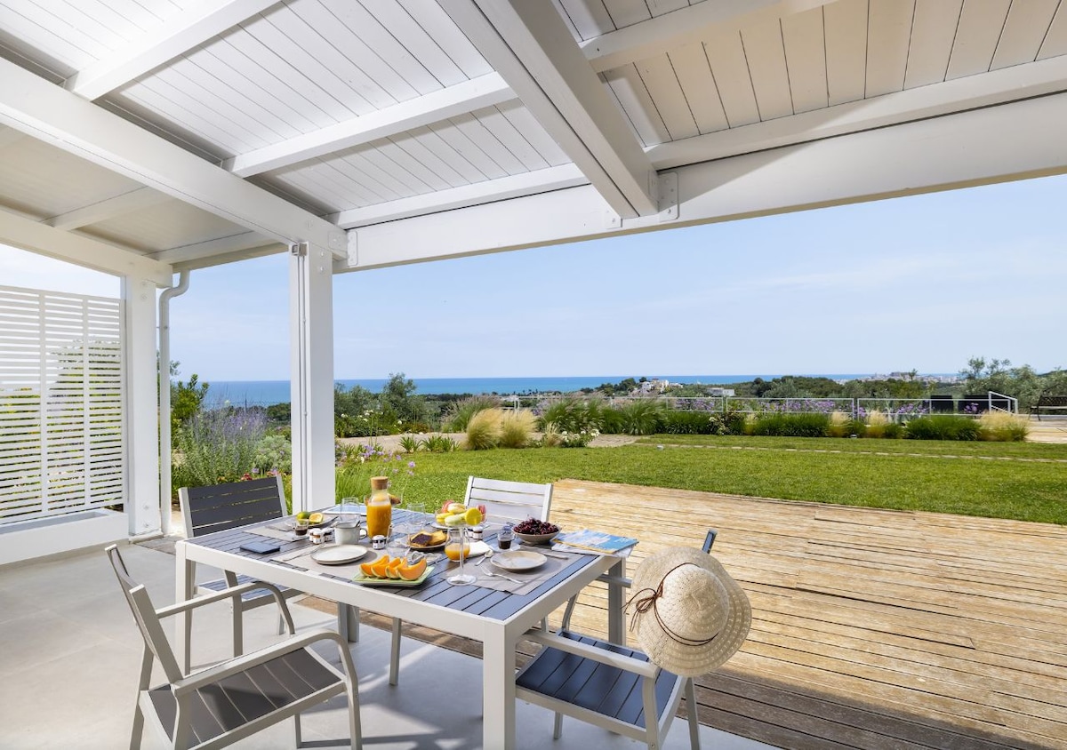 An outdoor dining area is set beneath a covered porch, featuring a wooden table adorned with a spread of fresh fruit, pastries, and beverages. The space opens to a lush garden and a distant view of the sea, creating a serene environment for meals.