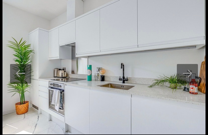 A modern kitchen features sleek white cabinetry and a spacious countertop. Stainless steel appliances, including a stove and kettle, are positioned for convenience. A small potted plant adds a touch of greenery, enhancing the clean and minimalist design.