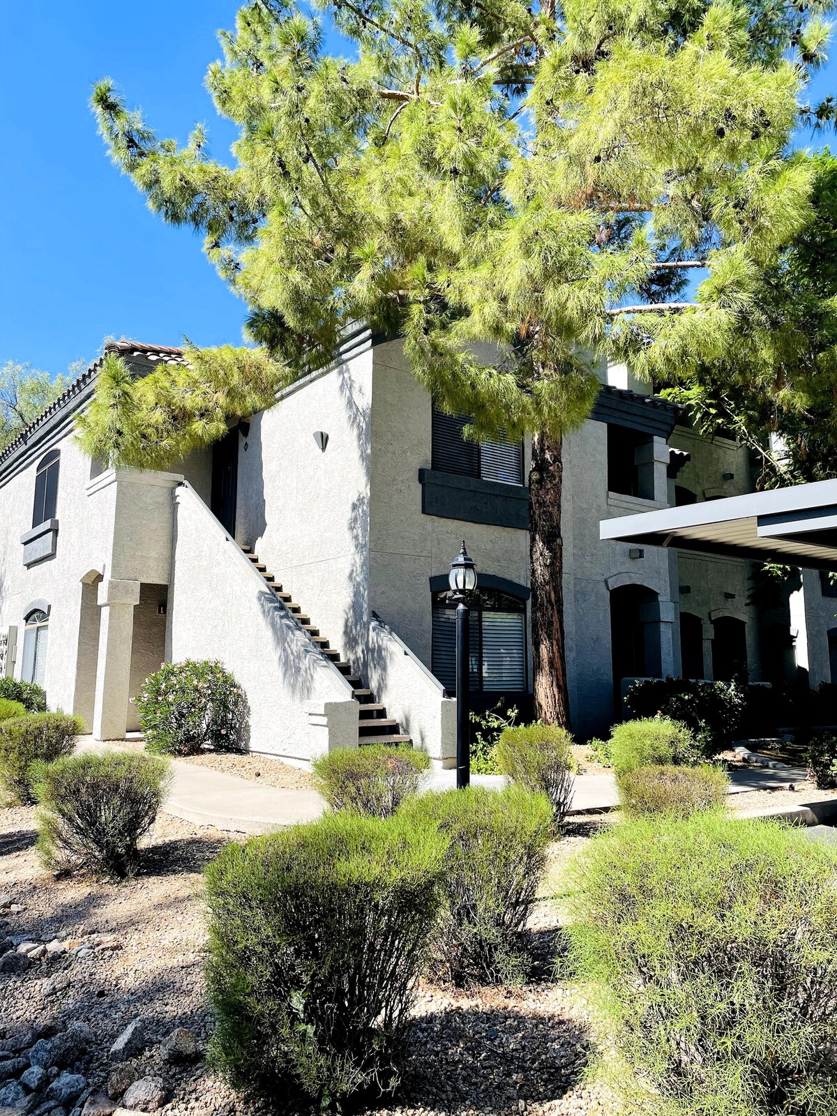 The exterior of a two-story building is surrounded by lush green landscaping, featuring a stairway leading to the upper units. Tall trees provide shade, while neatly trimmed bushes line the pathway. A lamp post adds a welcoming touch to the entrance area.