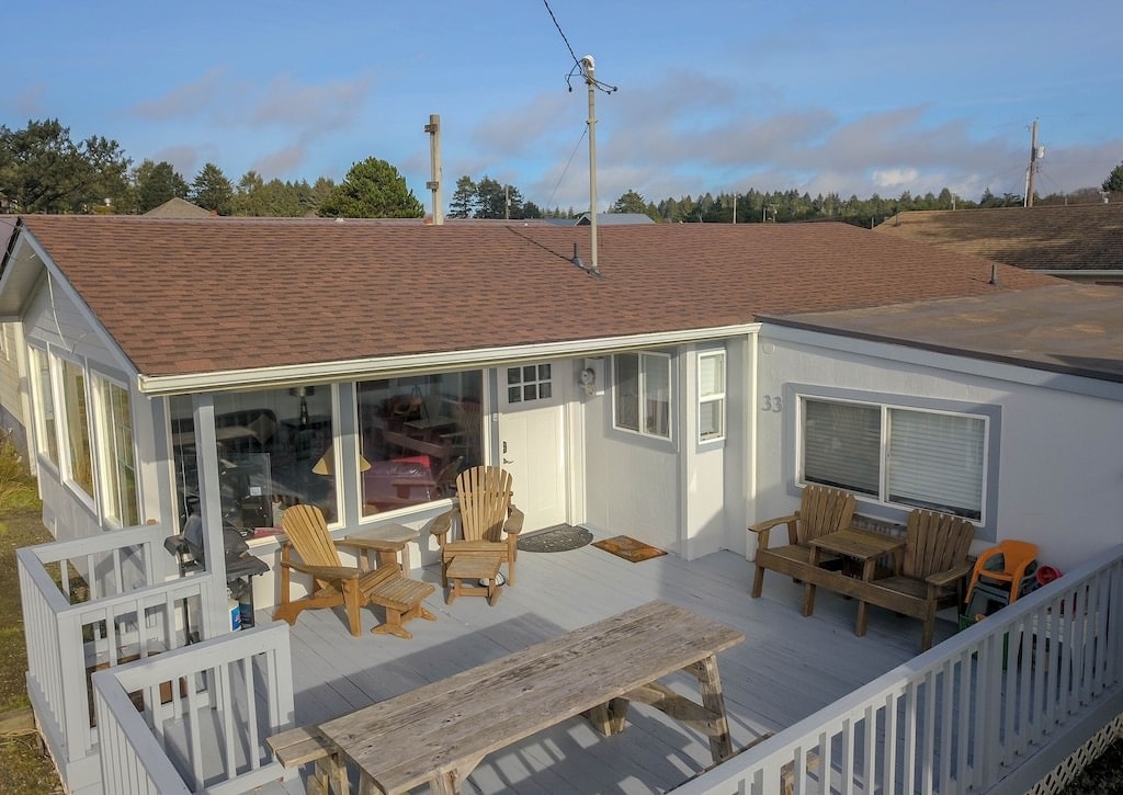 An outdoor deck area is featured, with a wooden table and several Adirondack chairs arranged for seating. A large window offers glimpses of the interior, while the surrounding landscape includes trees and open space in the background.