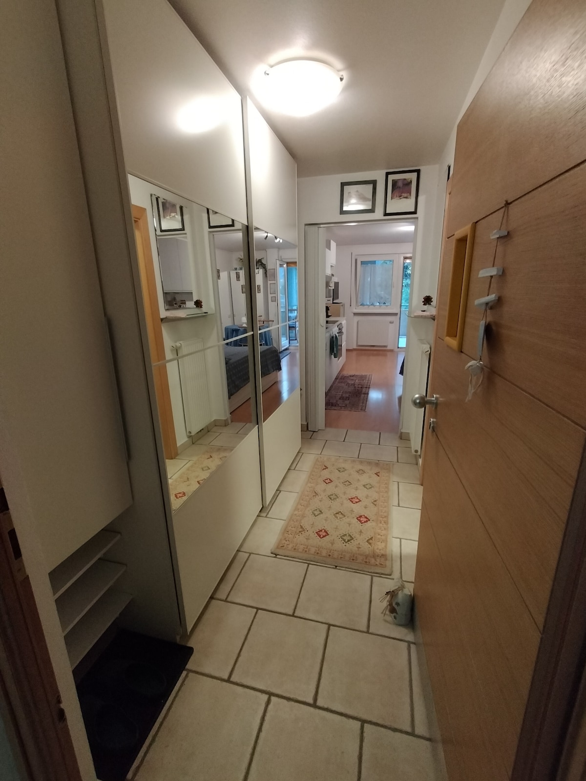 The entryway features a tiled floor and mirrored wardrobes along one side, creating a sense of space. A doormat is placed at the entrance, while natural light is visible through the doorway leading into the living area. Framed pictures are displayed on the wall.