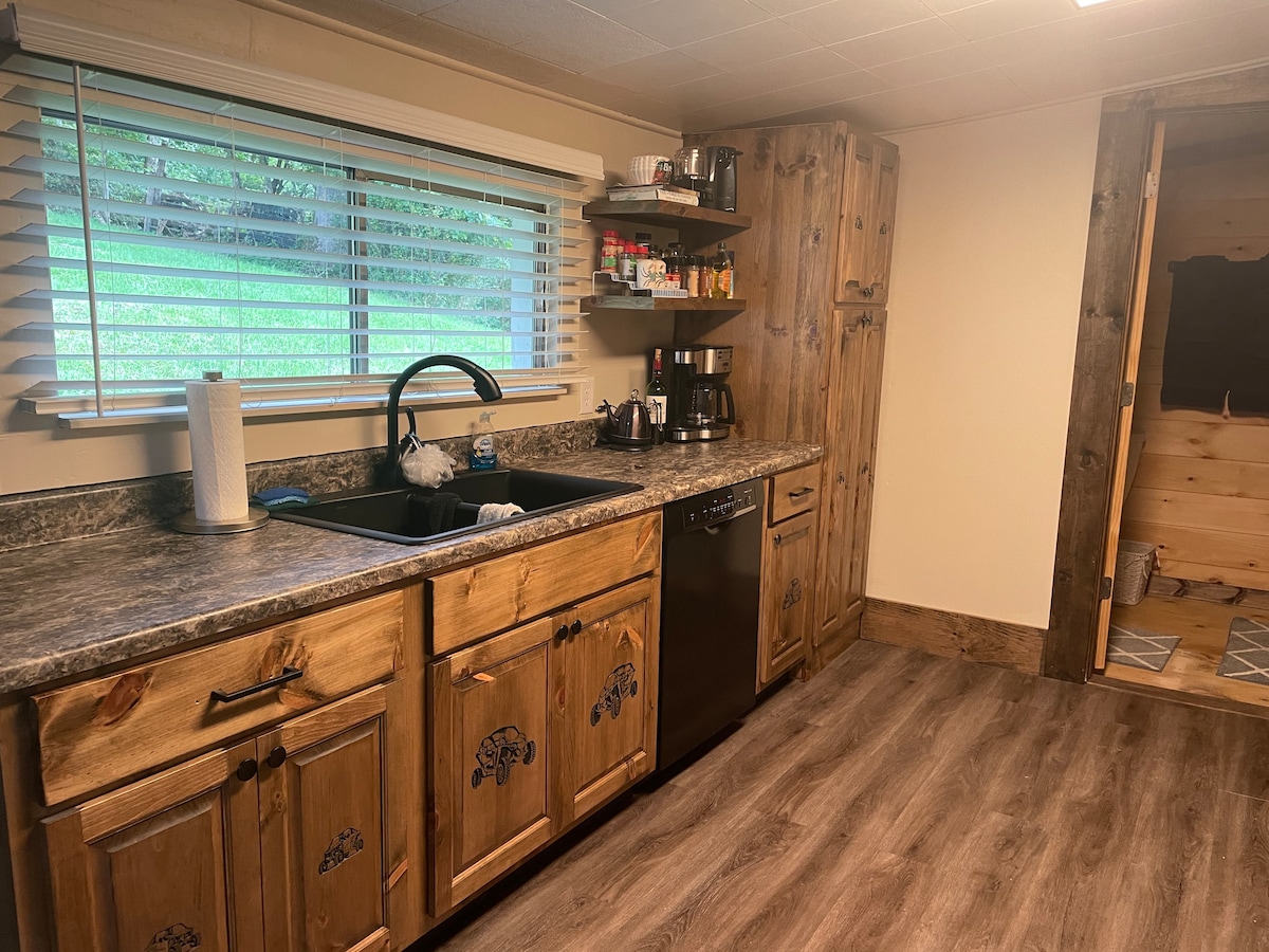 A fully equipped kitchen features wooden cabinetry with decorative accents, a modern sink, and stainless-steel appliances. Natural light floods the space through window blinds, highlighting the spacious countertop and organized shelves stocked with kitchen essentials.