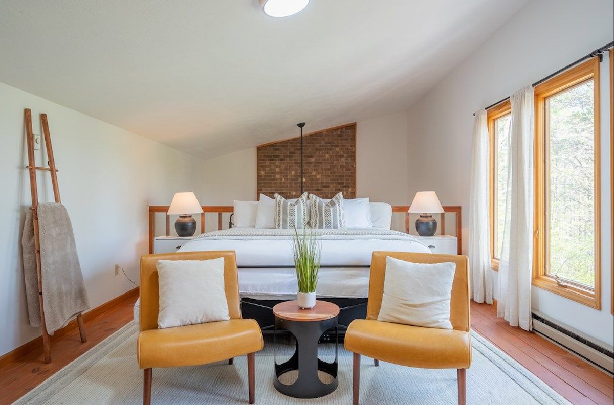 A spacious loft bedroom features a king bed with crisp white bedding, complemented by modern bedside lamps. Two inviting orange chairs are positioned around a circular table, while large windows let in ample natural light, enhancing the serene atmosphere of the space.