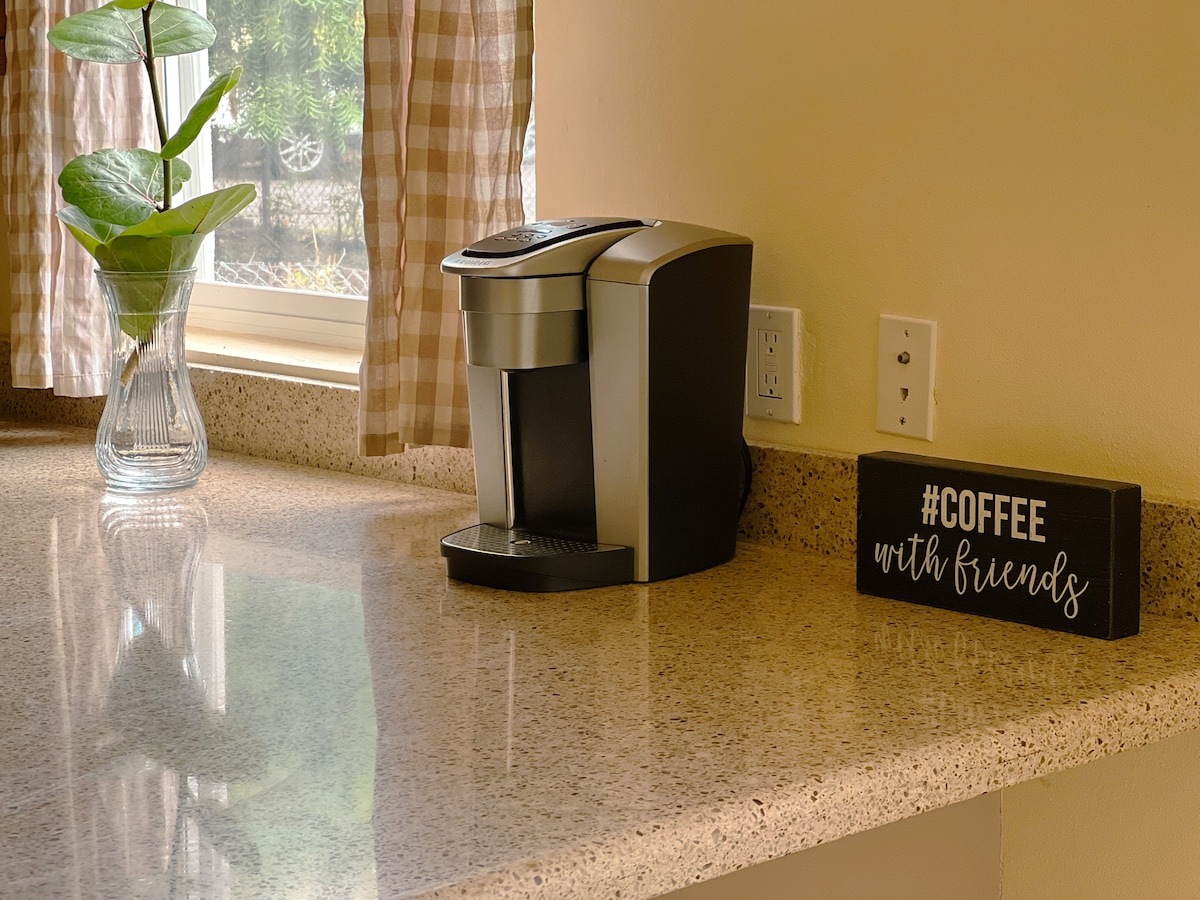 A sleek coffee maker sits on a smooth countertop, accompanied by a decorative sign reading '#COFFEE with friends'. A decorative vase with a green plant adds a touch of freshness, while the window provides natural light through soft, checked curtains.