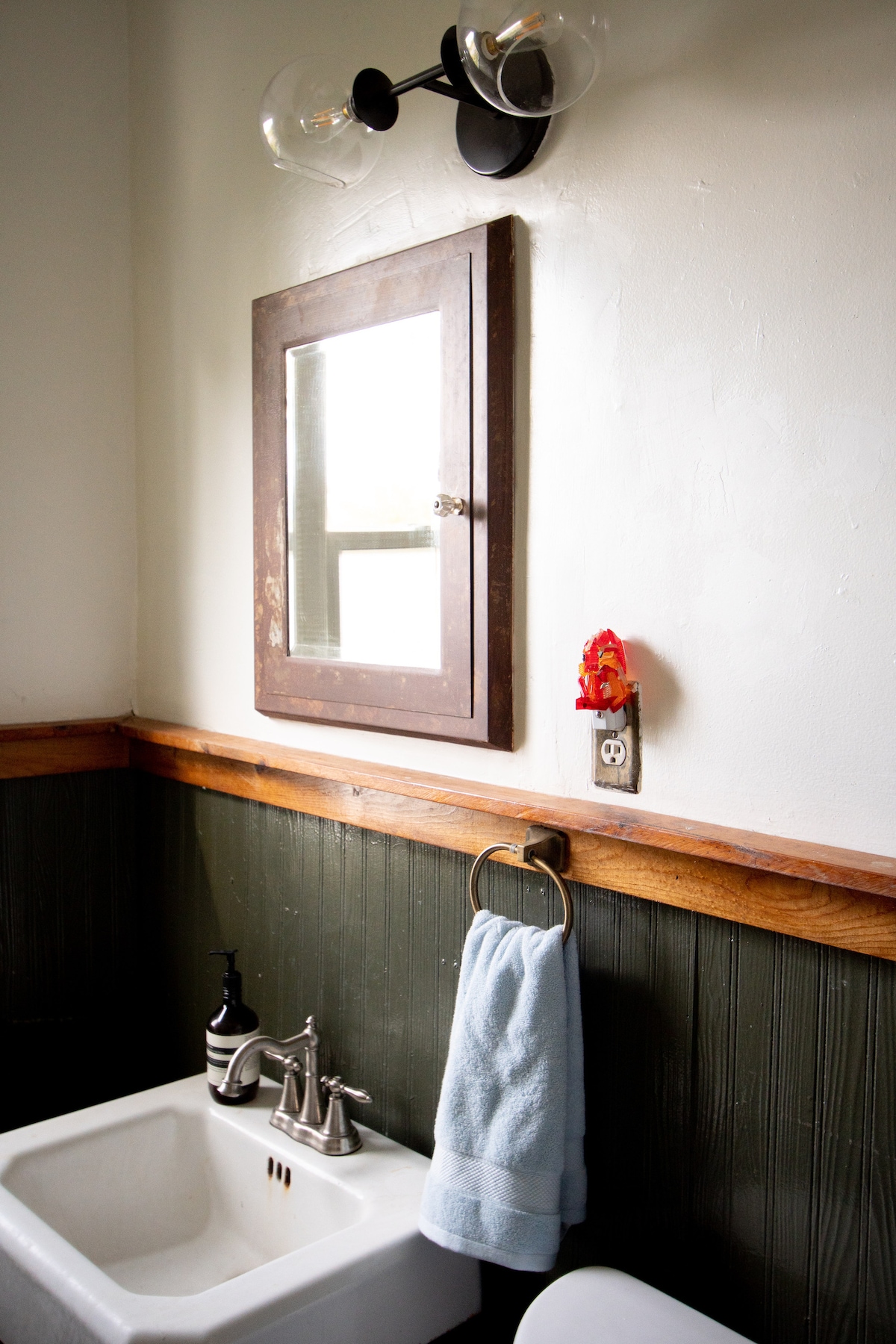 A bathroom is revealed featuring a vintage-style pedestal sink with modern fixtures. A wall-mounted mirror is framed in dark wood above the sink. Natural light enters through a nearby window, which is adjacent to a decorative wall element. A towel hangs from a ring beside the sink.